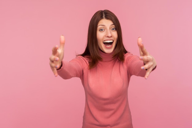 Amable mujer morena positiva en suéter rosa estirando las manos listas para abrazar mirando a la cámara con abrazos libres de felicidad Foto de estudio interior aislada sobre fondo rosa