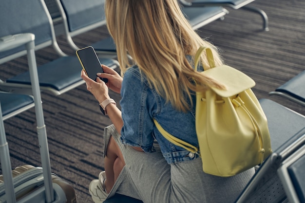 Amable mujer rubia manteniendo la mochila en la espalda mientras está sentada en la zona de salón y esperando la salida