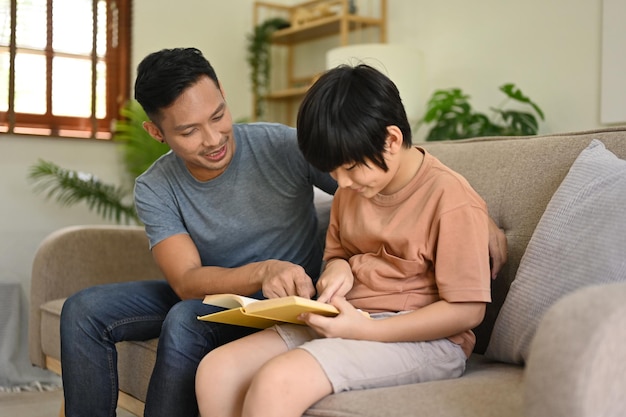 El amable padre asiático le está enseñando a su hijo a leer un libro o leyendo un libro juntos en un sofá