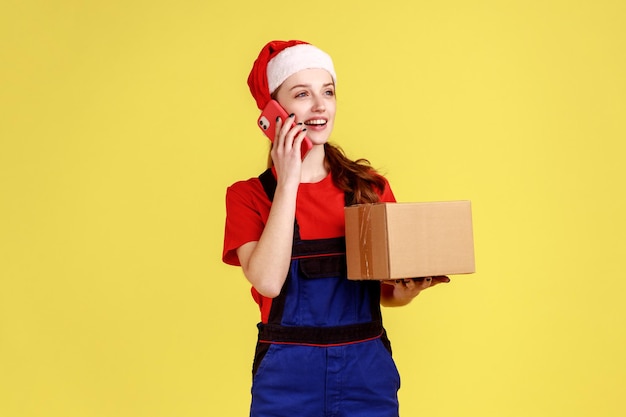 Amable repartidora hablando por teléfono celular con el cliente y aceptando el pedido sosteniendo una caja de cartón con un mono azul y un sombrero de santa claus Foto de estudio interior aislada sobre fondo amarillo