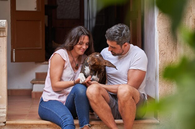 Amando a la mujer y al hombre con ropa informal sentados en las escaleras fuera de la casa y abrazando a un adorable perro juntos