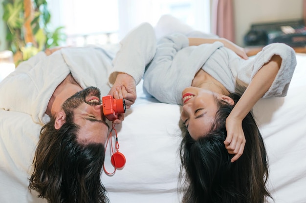 Amando o casal multiétnico em roupão de banho deitado na cama com a cabeça de cabeça para baixo brincando com uma câmera de brinquedo tirando fotografia