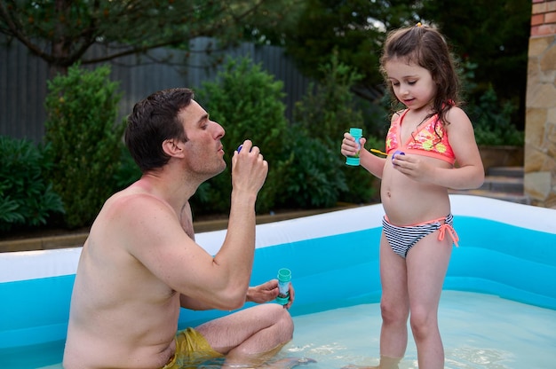 Amando a papá y su adorable bebé haciendo burbujas mientras se divierten en la piscina inflable en el jardín trasero