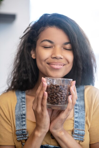 Amante del café. Alegremente sonriente joven con los ojos cerrados con un vaso de granos de café cerca de la cara