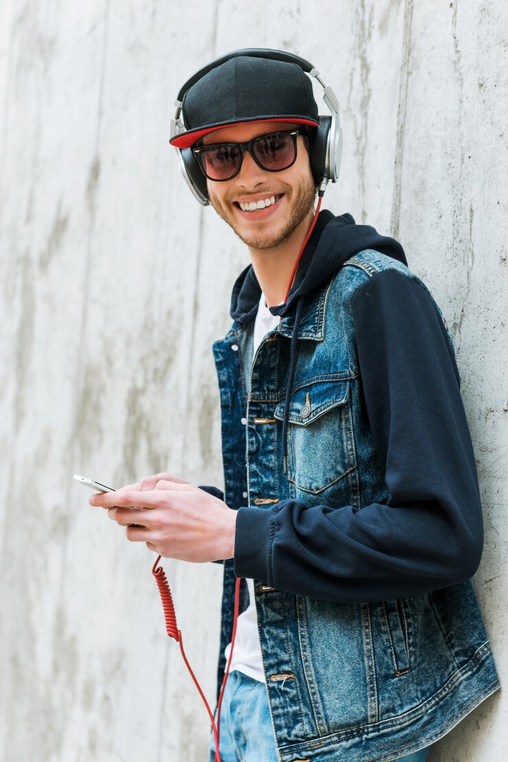 Amante da música. Jovem feliz em fones de ouvido, segurando um telefone celular enquanto se inclina para a parede de concreto