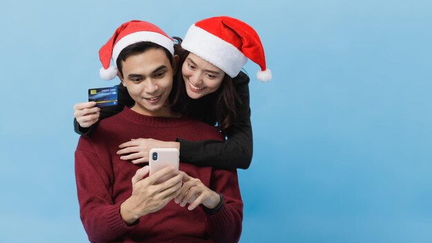 Amante do jovem casal asiático com chapéu de papai noel. compras on-line com cartão de crédito e smartphone isolado no fundo azul em um tiro de estúdio. celebração de natal do conceito de amante.