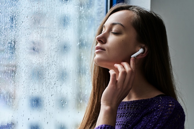 Amante de la música joven con los ojos cerrados en auriculares inalámbricos disfruta y escucha música relajante y relajante mientras está de pie junto a la ventana con gotas de lluvia en un clima lluvioso de otoño