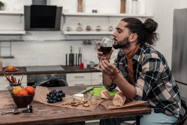 Amante del vino. Agradable joven disfrutando de su vino tinto mientras está sentado en la cocina