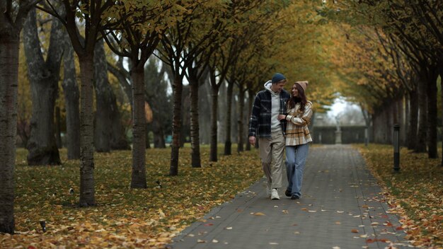 Amantes alegres caminhando felizes ao longo de um beco em um parque de folhagem Um belo casal de jovens se abraçando beijando ao ar livre Uma garota e um cara elegantes se divertindo juntos na natureza