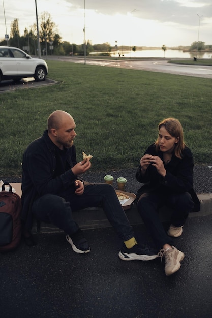 Amantes comem pizza sentado no gramado durante o pôr do sol