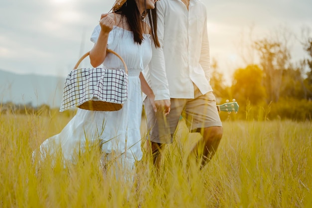 Los amantes felices están caminando y tomados de la mano en la pradera con ternura para encontrar un asiento para relajarse. Un hombre con un ukelele y una mujer con una caja de picnic.