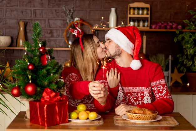 Amantes felices un hombre con una niña en suéteres rojos en la cocina con un árbol de Navidad y una bengala en casa celebran el año nuevo o la Navidad regocijándose, abrazándose y besándose