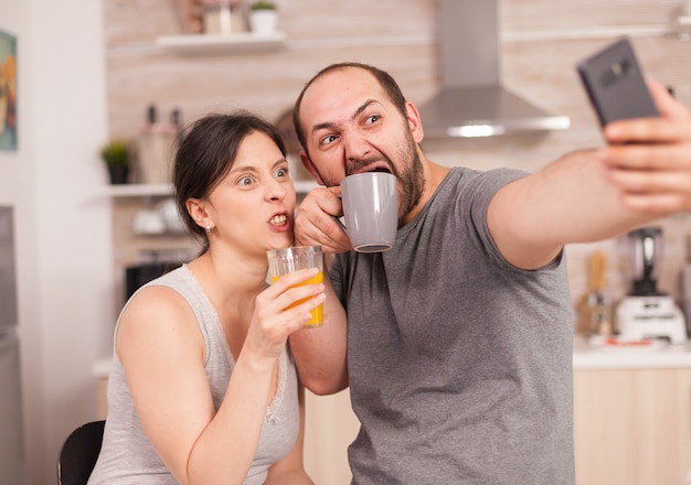 Amantes felices tomando selfies con el teléfono por la mañana bebiendo jugo de naranja y café. Marido y mujer casados alegres haciendo muecas mientras toman una foto durante el desayuno en la cocina.