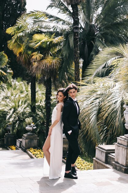 Foto amantes felizes a noiva e o noivo em roupas de casamento caminham entre plantas e palmeiras no velho parque