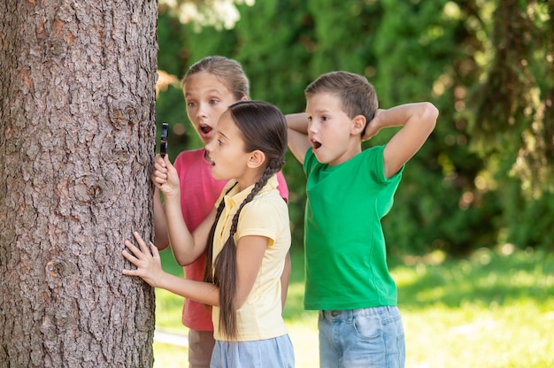 Amantes de la naturaleza. Chicas sorprendidas de pelo largo con la boca abierta con lupa y un niño de pie cerca de un árbol en el parque verde