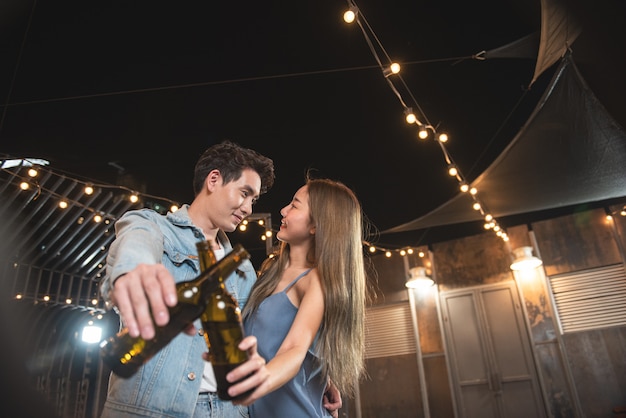 Foto los amantes de la pareja asiática joven se divierten bailando y bebiendo en la fiesta nocturna en la azotea del club nocturno con la botella de cerveza y el contacto visual coqueteando en la fiesta de pareja.