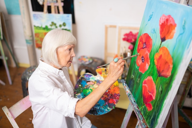 Amapolas sobre lienzo. Mujer jubilada con talento de pelo rubio pintando amapolas rojas sobre lienzo