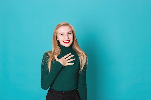 Amased schöne blonde Frau, die auf einer tragenden Strickjacke des blauen Hintergrundes steht