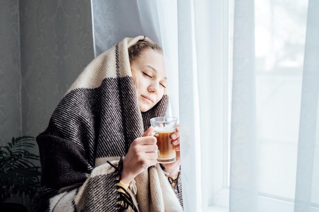 Ambiente acogedor bebé hace frío afuera Mujer joven de pie cerca de la ventana con plaid bebiendo café o té mirando afuera relajándose en su sala de estar