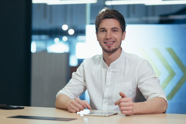 Ambiente de trabalho retrato de um jovem empresário masculino bonito sentado em uma mesa no escritório trabalhando em um laptop tablet celular ganhos funciona ele olha para a câmera sorri