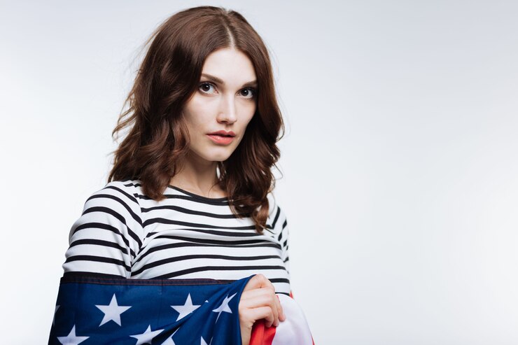 Foto ame meu país. linda mulher de cabelos castanhos com uma blusa listrada posando em um fundo cinza enquanto é enrolada em uma bandeira americana