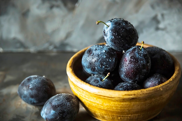 Ameixas azuis maduras em uma tigela de barro sobre uma mesa cinza. Conceito de frutas sazonais de verão