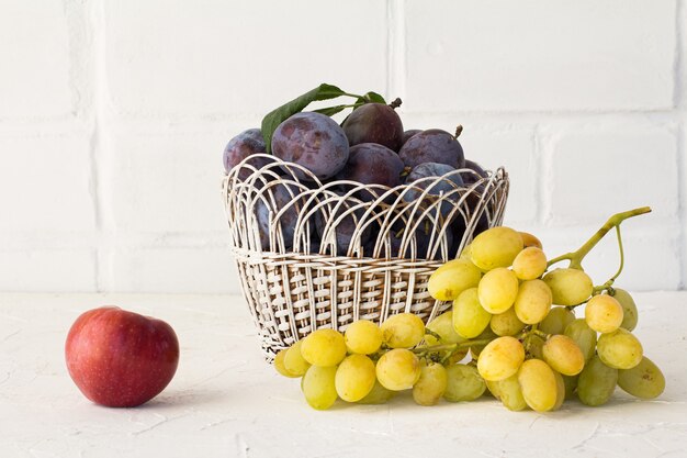 Ameixas maduras em uma cesta de vime, uma maçã e um cacho de uvas verdes maduras em um fundo de tijolos brancos