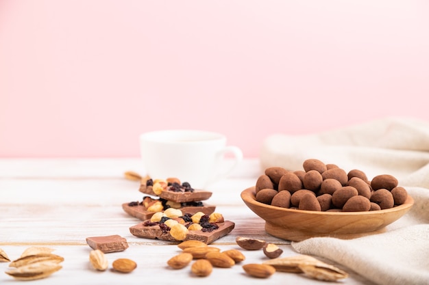 Amêndoa em drageias de chocolate em placa de madeira e uma xícara de café sobre fundo branco e rosa e têxteis de linho. Vista lateral, cópia espaço, foco seletivo.