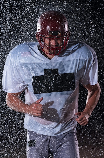 American Football Athlet Warrior, der auf einem Feld steht, hält seinen Helm und ist spielbereit. Spieler, der sich darauf vorbereitet, zu laufen, anzugreifen und einen Touchdown zu erzielen. Regnerische Nacht mit dramatischem Blendenfleck und Regentropfen. Hoch