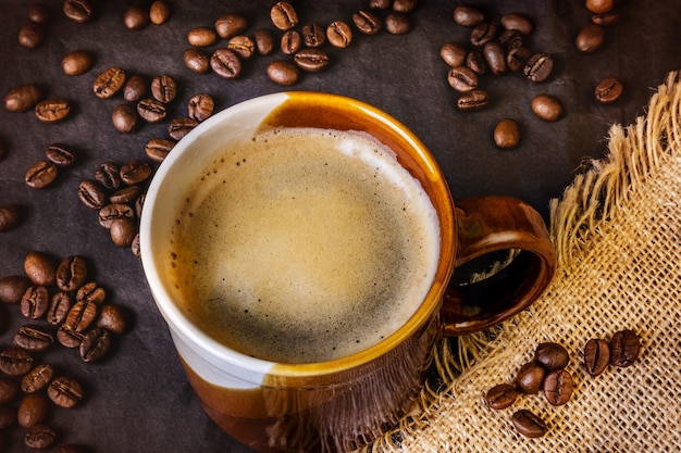 Americano na caneca branca e marrom em um escuro. Serapilheira fica nas proximidades, grãos de café estão espalhados