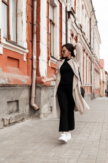 Amerikanische junge Frau in stilvoller Sonnenbrille in modischer Frühlingskleidung in weißen Schuhen mit Handtasche geht auf der Straße in der Nähe des Vintage-Gebäudes. Schönes Mädchen Hipster im trendigen Freizeitkleid reist in der Stadt.