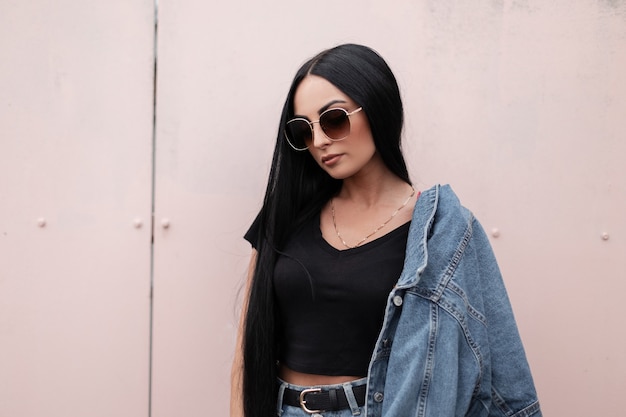 Amerikanische junge Hipster-Frau in stilvoller Sonnenbrille in einem schwarzen Oberteil in einer modischen blauen Jeansjacke in einem trendigen Rock steht nahe einer Wand in der Stadt im Freien. Ziemlich attraktives Mädchenmodel.