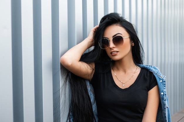 Amerikanische modische junge Frau mit luxuriösem langem schwarzen Haar im stilvollen T-Shirt in einer Sonnenbrille in einer Vintagen Jeansjacke, die nahe einer metallischen silbernen Wand in der Stadt aufwirft. Attraktives Hipster-Mädchen.