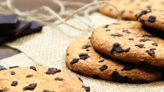 Amerikanische Schokoladenkekse auf einem braunen Holztisch und auf einer Leinenserviette Nahaufnahme. Traditioneller abgerundeter knuspriger Teig mit Schokoladenstückchen. Bäckerei. Leckeres Dessert, Gebäck. Ländliches Stillleben.