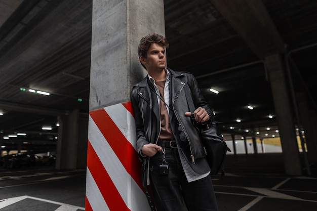 Amerikanischer junger Mann in einer modischen schwarzen Lederjacke in einem klassischen Hemd mit einem Vintage-Rucksack mit einer Frisur, die in der Nähe einer Säule mit einer rot-weißen Linie auf einem Stadtparkplatz posiert. Urban cooler trendiger Typ