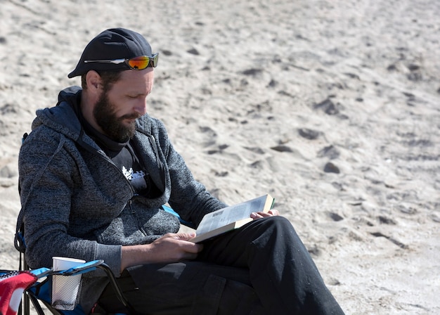 Amerikanischer Mann, der sich bei Sonnenuntergang auf einem Buch am Strand entspannt. Russischer Mann liest ein Buch im Liegestuhl mit Blick auf das Meer. gut aussehender kaukasischer bärtiger Mann mit Sonnenbrille, Mütze und Kapuzenjacke