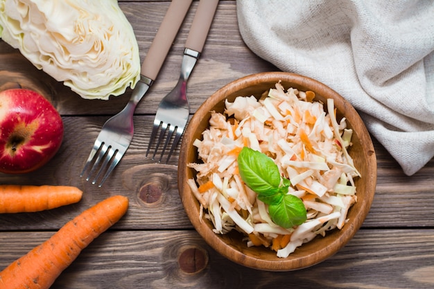 Amerikanischer verzehrfertiger Krautsalat-Salat aus Kohl, Sellerie, Karotten und Äpfeln mit Basilikumblättern in einem Holzteller und Zutaten zum Kochen auf einem Holztisch. Gesundes und essendes Konzept.