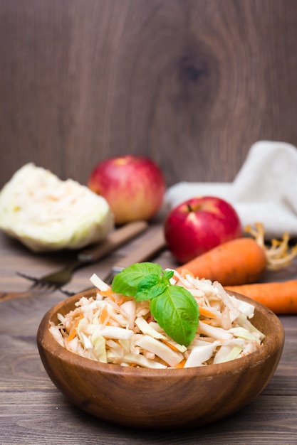 Amerikanischer verzehrfertiger Krautsalat-Salat aus Kohl, Sellerie, Karotten und Äpfeln mit Basilikumblättern in einem Holzteller und Zutaten zum Kochen auf einem Holztisch.