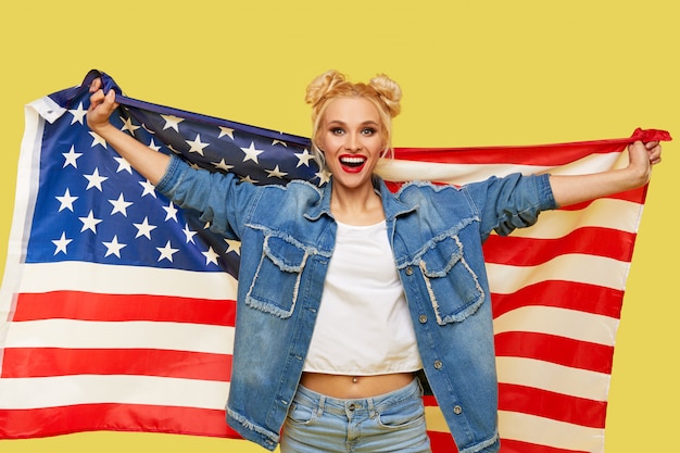 Amerikanisches Mädchen. Glückliche junge Frau in Jeanskleidung, die USA-Flagge lokalisiert auf gelbem Hintergrund hält