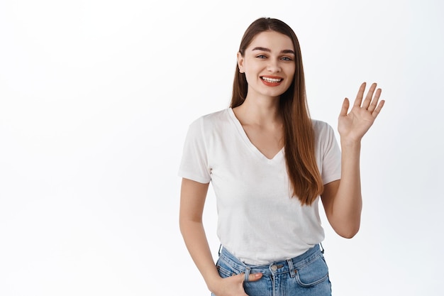 Foto amigable chica saludando con la mano y saludar, saludar, dar la bienvenida a los invitados, hacer un gesto de hola, una sonrisa positiva, de pie sobre fondo blanco.