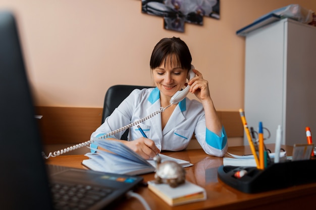 Una amigable doctora se sienta en su oficina y habla por teléfono con uno de los pacientes y escribe algo en un cuaderno. El doctor esta trabajando. El concepto de medicina.