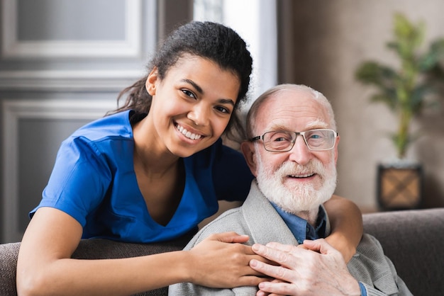 Foto amigable médico general maduro comunicándose con un agradable paciente masculino de los años ochenta sentados juntos en el sofá sonriendo confiado joven médico dando ayuda psicológica a un anciano en visita a casa