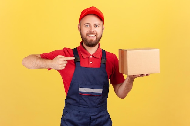 Amigable mensajero en uniforme apuntando a la caja de cartón entregando el paquete al cliente