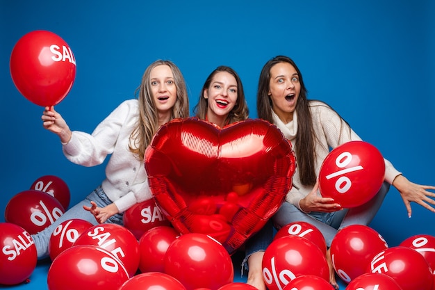 Amigas bonitas felices que se sientan con los globos rojos de la venta, aislados en la pared azul