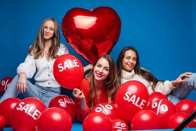 Foto amigas bonitas felices que se sientan con los globos rojos de la venta, aislados en la pared azul