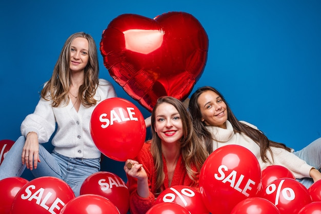 Foto amigas bonitas felices que se sientan con los globos rojos de la venta, aislados en la pared azul