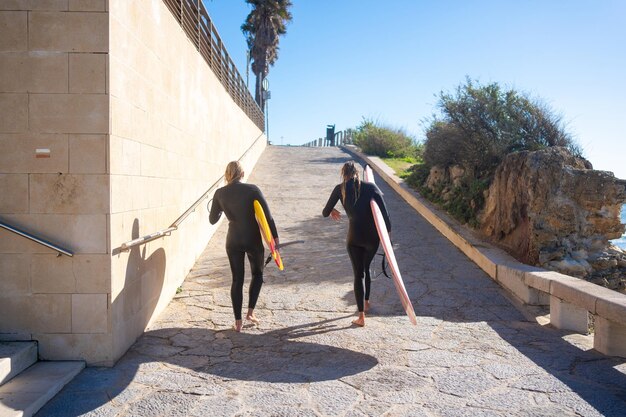 Amigas caminhando com pranchas de surf na passarela sob o céu azul