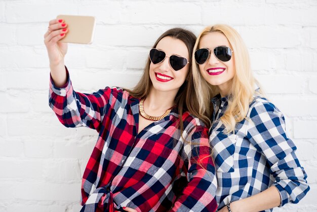 Amigas en camisas a cuadros haciendo retrato selfie con teléfono en la pared blanca de fondo