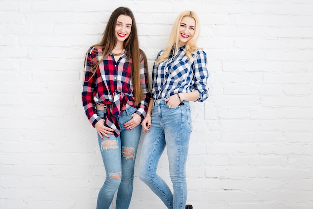 Amigas en camisas a cuadros y jeans parados juntos en el fondo de la pared blanca