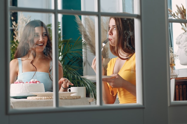 Amigas en casa sentados y sonriendo con pastel de cumpleaños blanco detrás de la puerta de cristal.